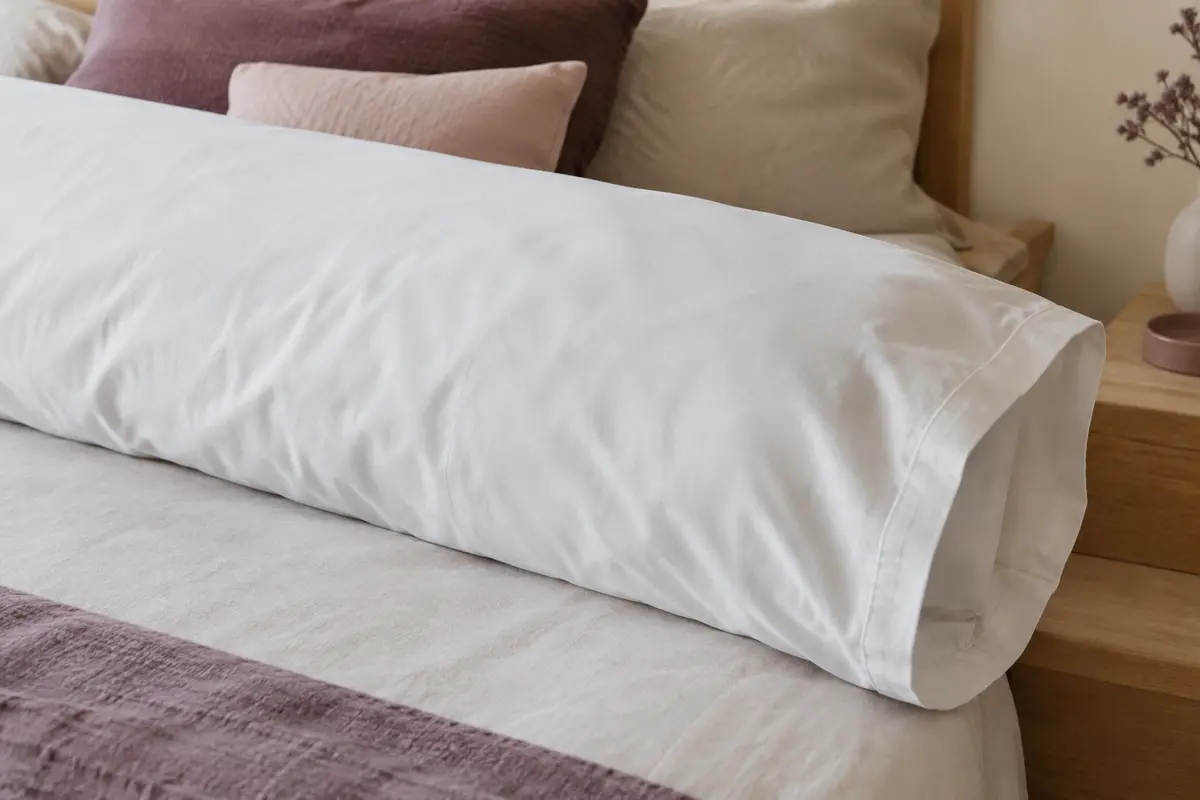 Taie de traversin blanche en coton percale posée sur un lit aux tons ivoire et prune doux.