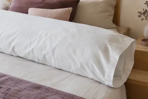 Taie de traversin blanche en coton percale posée sur un lit aux tons ivoire et prune doux.