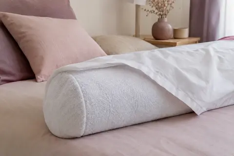 Protège-traversin blanc en éponge installé sur un traversin, dans une chambre aux tons ivoire et prune doux.