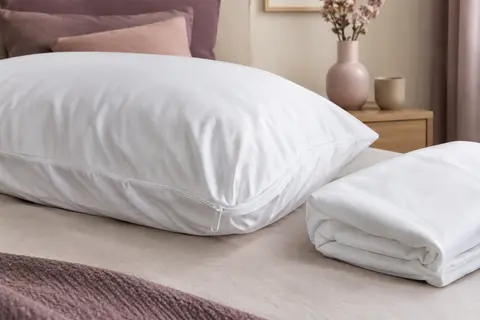 Protège-oreiller blanc à fermeture zippée posé sur un oreiller, avec un second exemplaire plié à côté.