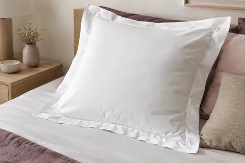 Taie d’oreiller carrée blanche en coton avec volant plat, posée sur un lit dans une chambre aux tons doux.
